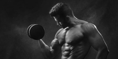 Powerful black and white portrait of a shirtless bodybuilder lifting dumbbells in the gym.